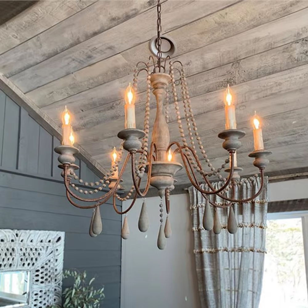 A French country retro style 6-head chandelier, featuring a weathered solid wood main frame paired with iron lamp arms, wooden bead chains and teardrop pendants, using candle-shaped bulbs. It emits a warm and soft glow when lit, combining decorative appeal with practical illumination, ideal for creating a cozy and retro space atmosphere.