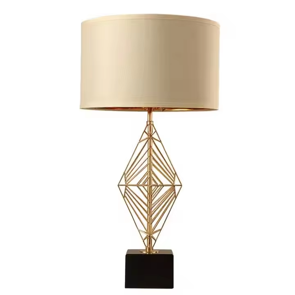 This is a luxury geometric diamond-shaped table lamp with a golden geometric hollow diamond bracket and a beige fabric lampshade. It features a simple and elegant design with excellent texture. It is used for local desktop lighting and decoration, creating a luxurious and warm ambiance. It is suitable for luxury-style home bedroom bedside tables, high-end hotel guest rooms, modern soft decoration display spaces and other places.