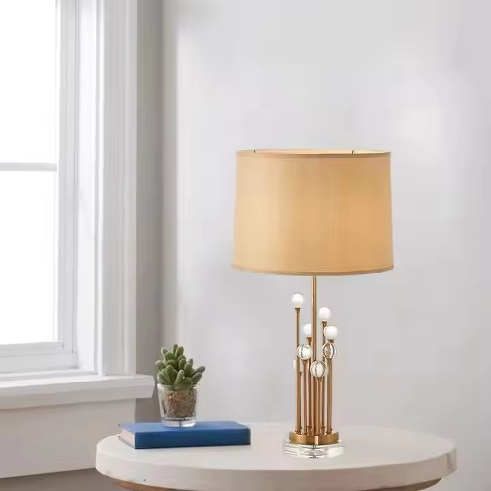 This is a modern luxury-style table lamp with a golden metal stand, multiple spherical decorations, and a beige fabric lampshade that transmits light softly. It has an exquisite and layered design. It is used for local desktop lighting and decoration, combining practicality and aesthetics. It is suitable for modern home living room side tables, bedroom bedside tables, as well as high-end hotel guest rooms, luxury cafes and other places.