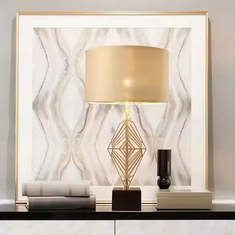 This is a luxury geometric diamond-shaped table lamp with a golden geometric hollow diamond bracket and a beige fabric lampshade. It features a simple and elegant design with excellent texture. It is used for local desktop lighting and decoration, creating a luxurious and warm ambiance. It is suitable for luxury-style home bedroom bedside tables, high-end hotel guest rooms, modern soft decoration display spaces and other places.