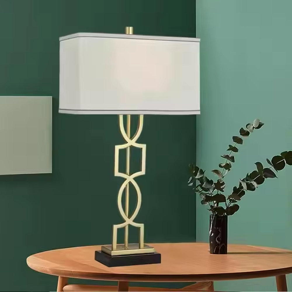 This is a luxury-style table lamp with a golden geometric hollow bracket and a white fabric square lampshade. It features a simple and elegant design with excellent texture. It is used for local desktop lighting and decoration, creating a luxurious and warm ambiance. It is suitable for luxury-style home bedroom bedside tables, high-end hotel guest rooms, modern office spaces and other places.