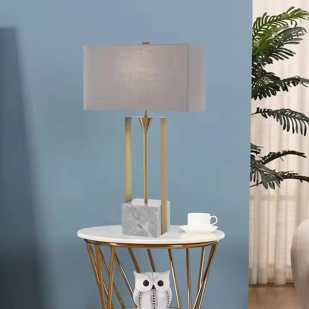 This is a modern minimalist table lamp with a golden metal frame, a marble base, and a gray fabric square lampshade, featuring a simple and elegant design. It is used for local desktop lighting and decoration, combining practicality and aesthetics. It is suitable for modern home living room side tables, bedroom bedside tables, high-end hotel guest rooms, luxury office spaces and other places.