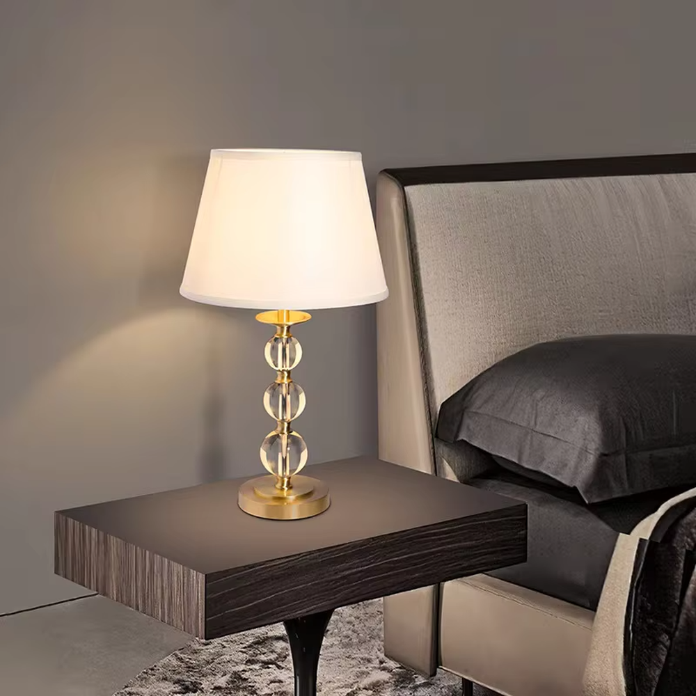 This is a luxury crystal ball table lamp with a golden metal frame, multi-layer transparent crystal balls, and a beige fabric lampshade, featuring a minimalist luxury design. It is used for local desktop lighting and decoration, creating a warm and luxurious ambiance. It is suitable for luxury-style home bedroom bedside tables, high-end hotel guest rooms, modern soft decoration display spaces and other places.