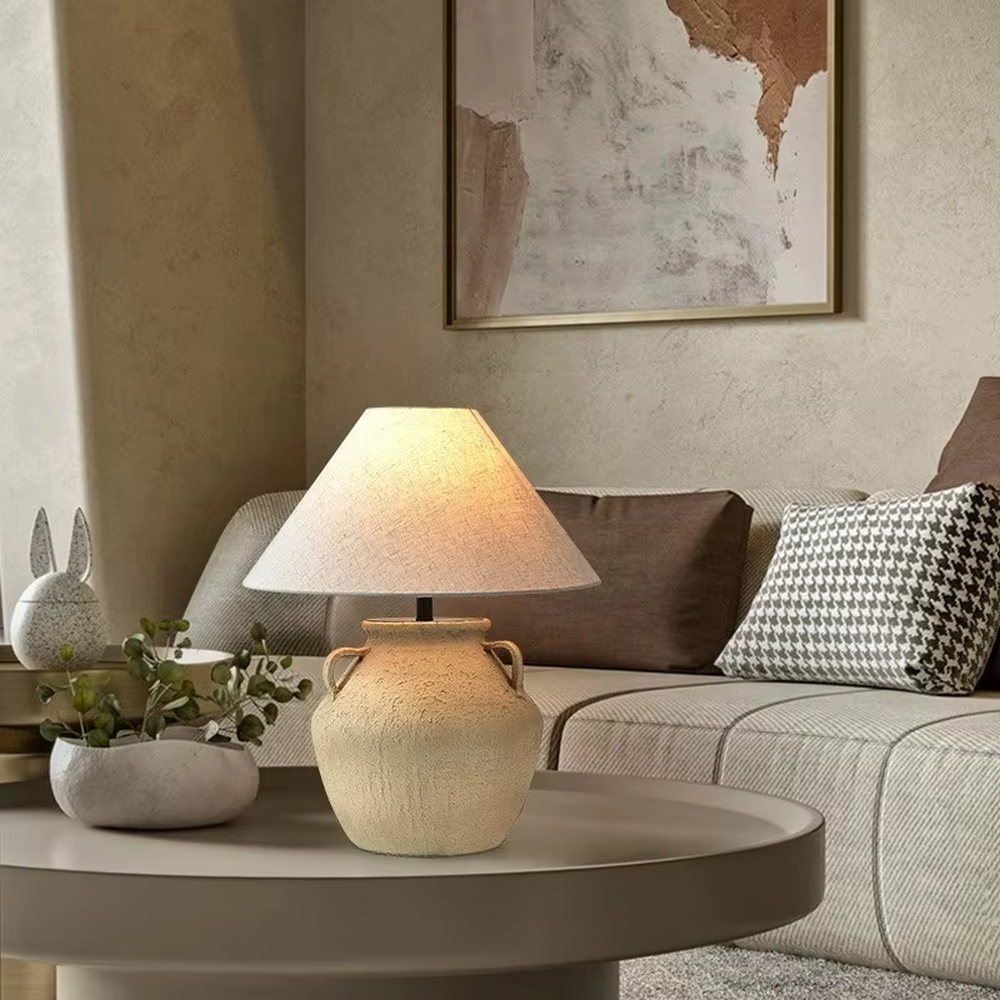 This is a retro clay pot - shaped table lamp with a beige textured clay pot base (with double handles) and a beige fabric slant lampshade, featuring a natural and rustic design with retro artistic sense. It is used for local desktop lighting and decoration, creating a warm retro atmosphere. It is suitable for wabi - sabi style home living rooms, retro - style homestays, art soft decoration display spaces and other places.