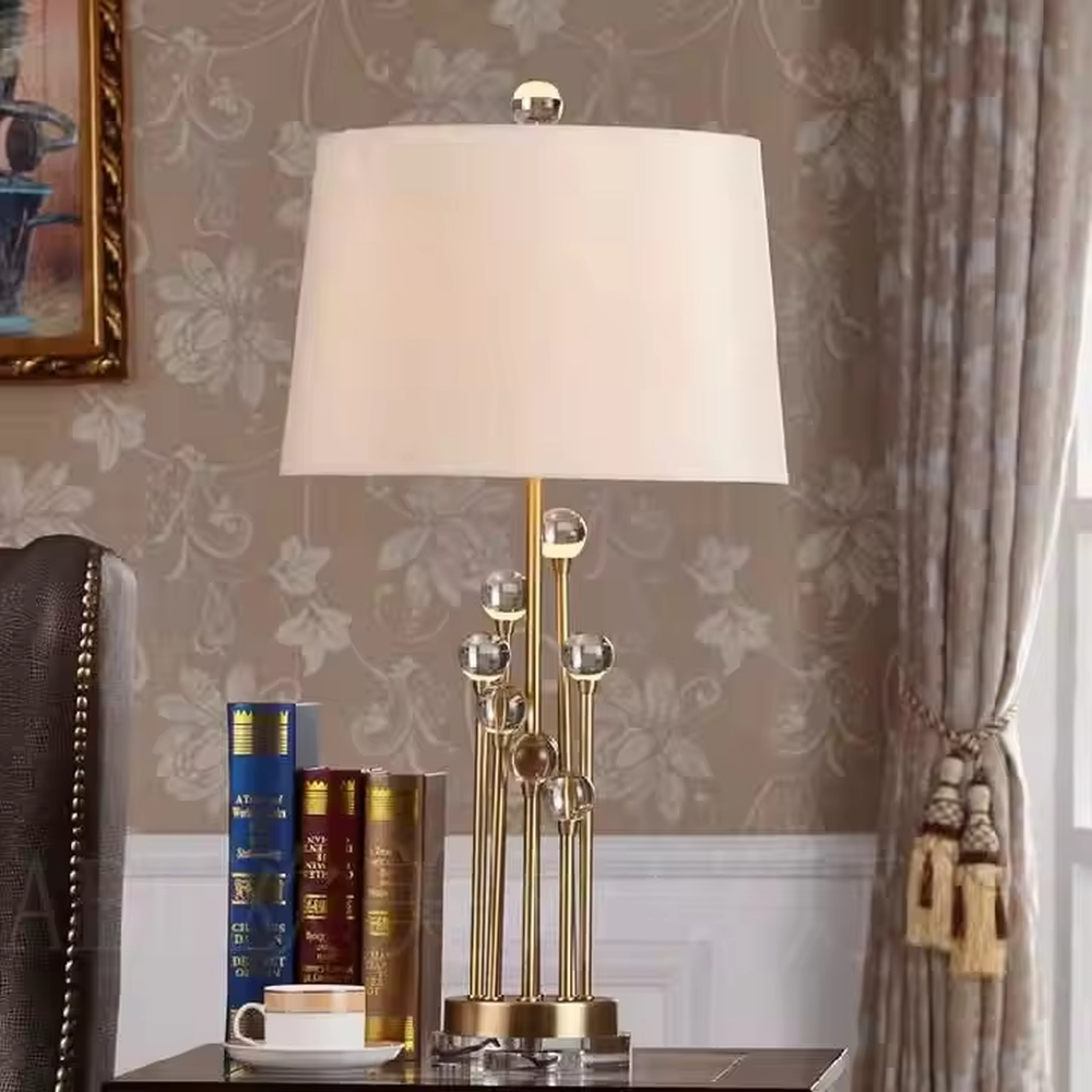 This is a modern luxury-style table lamp with a golden metal stand, multiple spherical decorations, and a beige fabric lampshade that transmits light softly. It has an exquisite and layered design. It is used for local desktop lighting and decoration, combining practicality and aesthetics. It is suitable for modern home living room side tables, bedroom bedside tables, as well as high-end hotel guest rooms, luxury cafes and other places.