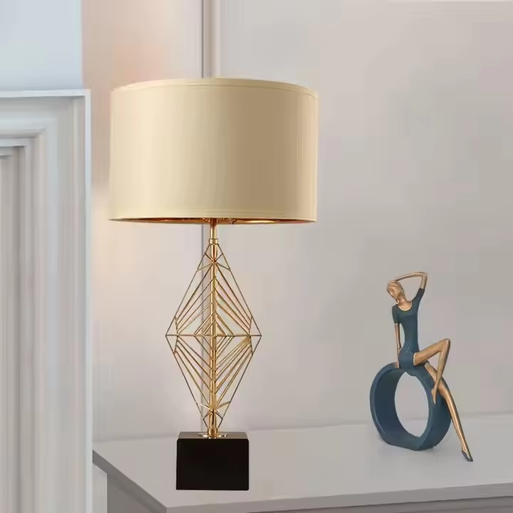 This is a luxury geometric diamond-shaped table lamp with a golden geometric hollow diamond bracket and a beige fabric lampshade. It features a simple and elegant design with excellent texture. It is used for local desktop lighting and decoration, creating a luxurious and warm ambiance. It is suitable for luxury-style home bedroom bedside tables, high-end hotel guest rooms, modern soft decoration display spaces and other places.