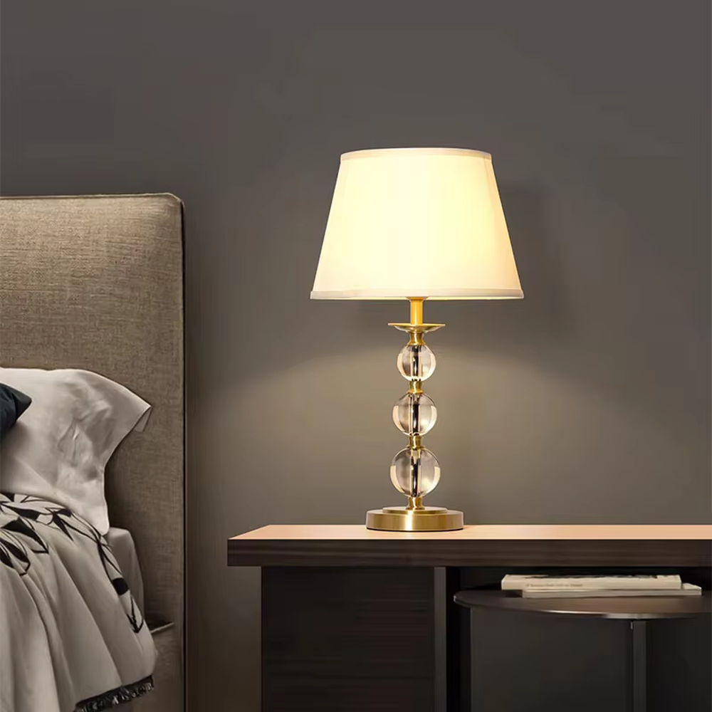 This is a luxury crystal ball table lamp with a golden metal frame, multi-layer transparent crystal balls, and a beige fabric lampshade, featuring a minimalist luxury design. It is used for local desktop lighting and decoration, creating a warm and luxurious ambiance. It is suitable for luxury-style home bedroom bedside tables, high-end hotel guest rooms, modern soft decoration display spaces and other places.