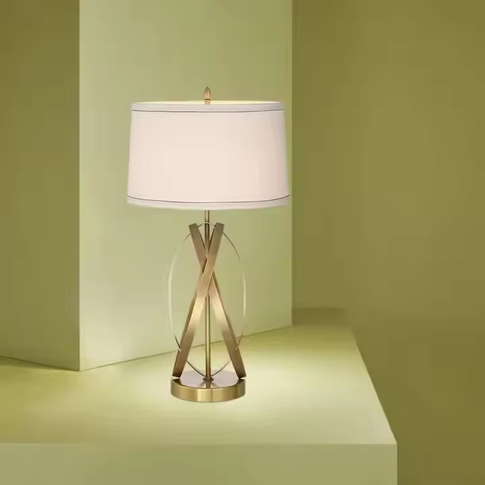 This is a luxury-style table lamp with golden crossed metal brackets and a beige fabric lampshade. It features a simple and elegant design with excellent texture. It is used for local desktop lighting and decoration, creating a luxurious and warm ambiance. It is suitable for luxury-style home bedroom bedside tables, high-end hotel guest rooms, modern office spaces and other places.