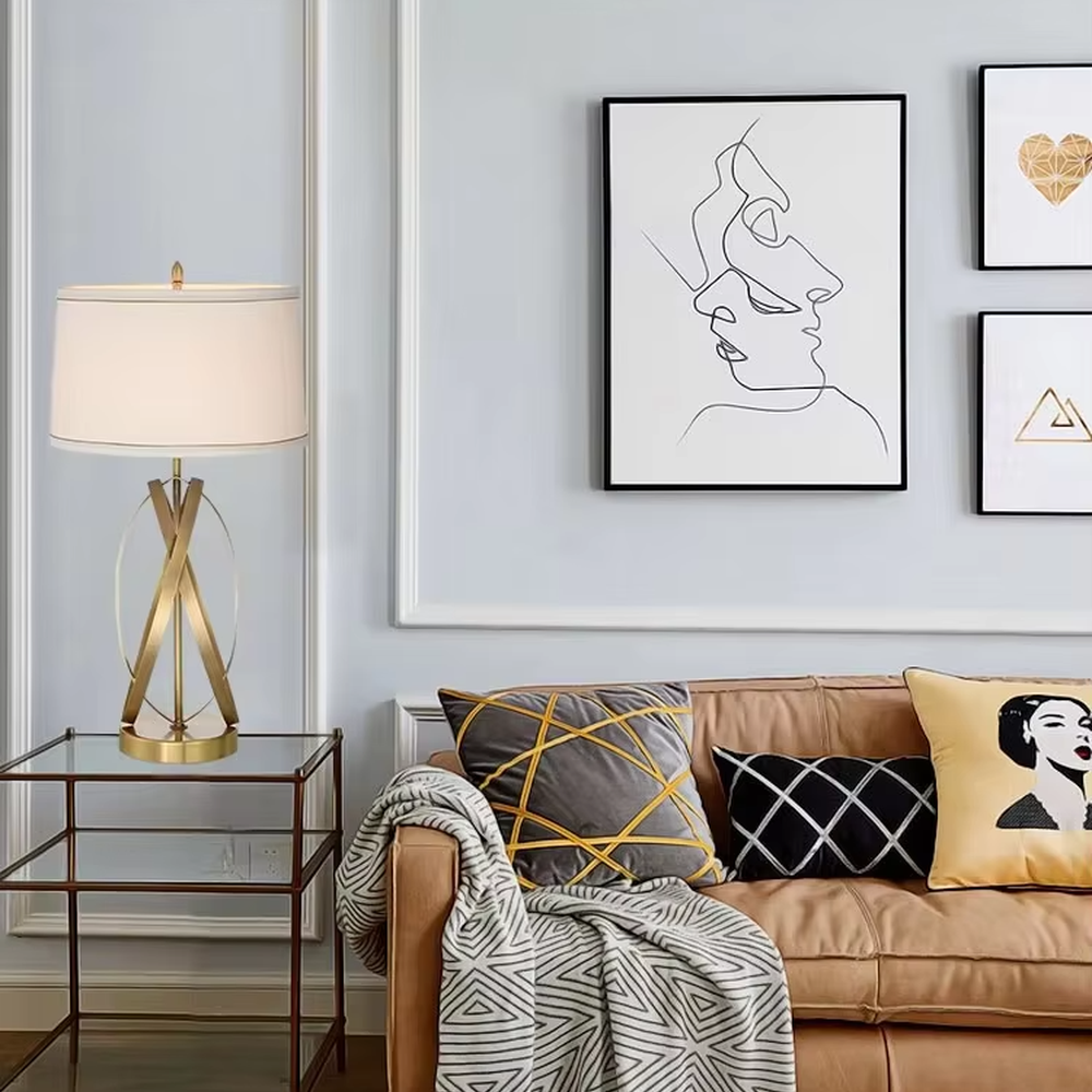 This is a luxury-style table lamp with golden crossed metal brackets and a beige fabric lampshade. It features a simple and elegant design with excellent texture. It is used for local desktop lighting and decoration, creating a luxurious and warm ambiance. It is suitable for luxury-style home bedroom bedside tables, high-end hotel guest rooms, modern office spaces and other places.