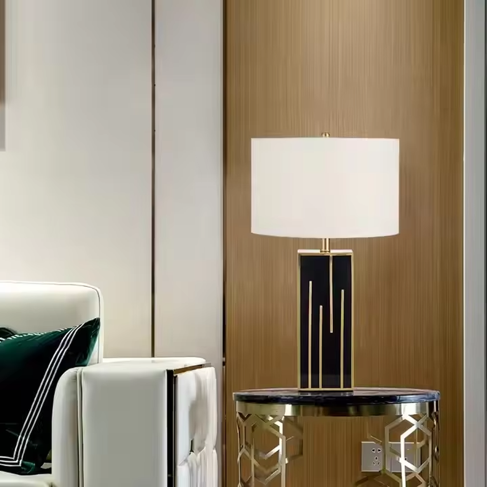 This is a luxury geometric - shaped table lamp with a black - gold geometric base (decorated with gold lines) and a white cylindrical fabric lampshade, featuring a minimalist luxury design with strong modern artistic sense. It is used for local desktop lighting and decoration, creating a luxurious and elegant atmosphere. It is suitable for luxury - style home bedroom bedside tables, high - end hotel guest rooms, modern soft decoration display spaces and other places.