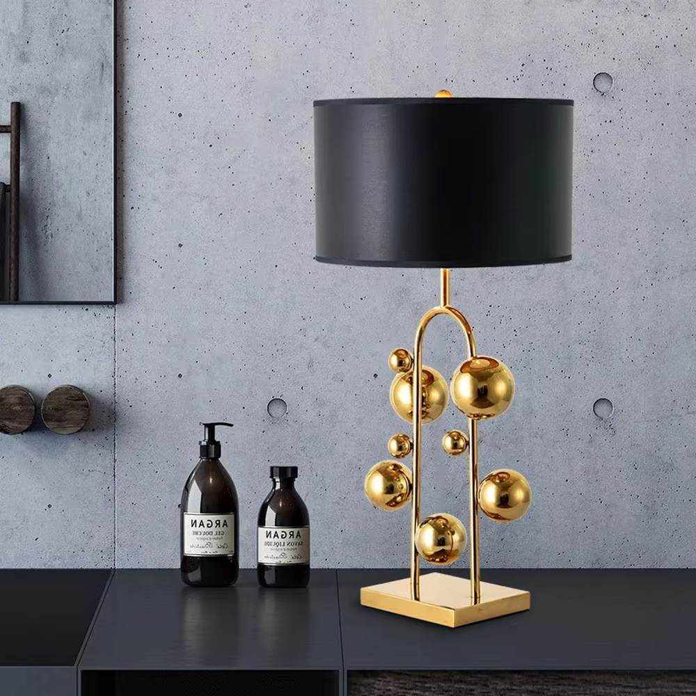 This is a light luxury modern-style golden ball table lamp, with a golden metal frame paired with multi-sized golden ball decorations, a black cylindrical lampshade + a golden square base, featuring a high-end and exquisite style, with soft and stylish lighting.