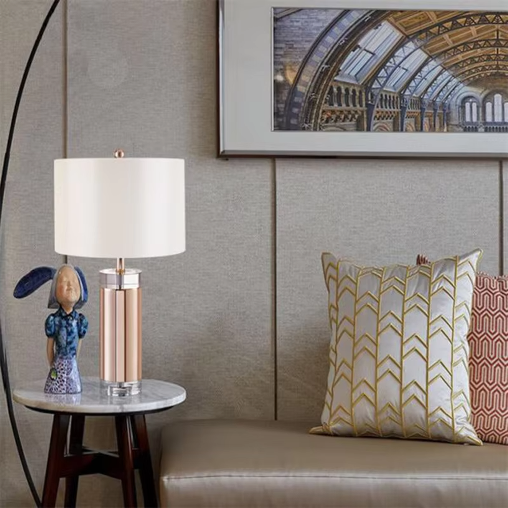 This is a luxury metal crystal table lamp, with a golden metal column, a transparent crystal base, and a white cylindrical fabric lampshade (with golden decoration on top), featuring a luxurious and elegant design with strong modern sense. It is used for local desktop lighting and decoration, creating a high-end luxury atmosphere. It is suitable for luxury-style home bedroom bedside tables, high-end hotel guest rooms, modern soft decoration display spaces and other places.