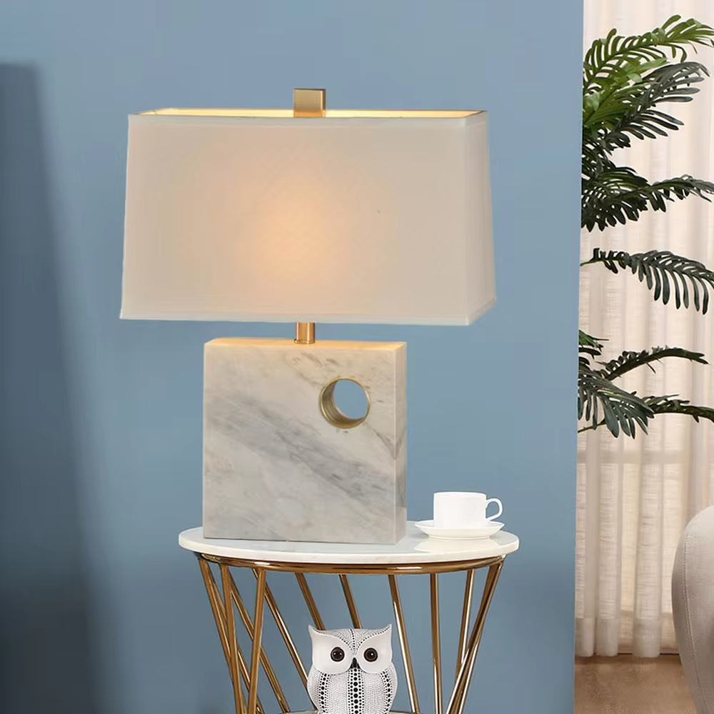 This is a light luxury elegant marble-based fabric table lamp, with a white marble base with golden ring decoration, paired with a beige fabric square lampshade and golden metal connectors. It features a minimalist light luxury design, with warm and soft lighting. It is used for local desktop lighting and decoration, creating a light luxury elegant atmosphere. Suitable for light luxury-style bedroom bedside tables, modern minimalist studies, high-end hotel guest rooms and other places.