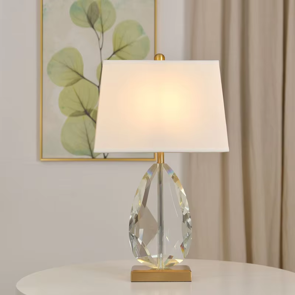 This is a light luxury exquisite water-drop shaped crystal table lamp, with a transparent cut water-drop crystal base paired with golden metal parts and a white square fabric lampshade. The design is simple and luxurious, with soft and elegant lighting. It is used for local desktop lighting and decoration, creating a light luxury exquisite atmosphere. Suitable for light luxury-style bedroom bedside tables, modern minimalist living room side tables, high-end hotel guest rooms and other places.
