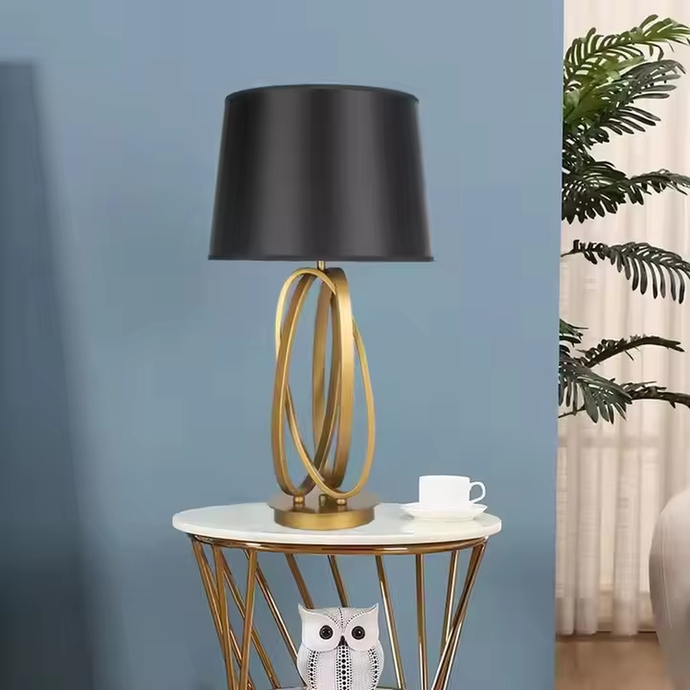 This is a modern luxury circular metal table lamp, with a golden interlaced circular metal base and a black fabric conical lampshade, featuring an artistic design and strong luxury texture. It is used for local desktop lighting and decoration, creating a high-end luxury atmosphere. It is suitable for luxury-style home bedroom bedside tables, modern minimalist living room side tables, high-end hotel guest rooms and other places.