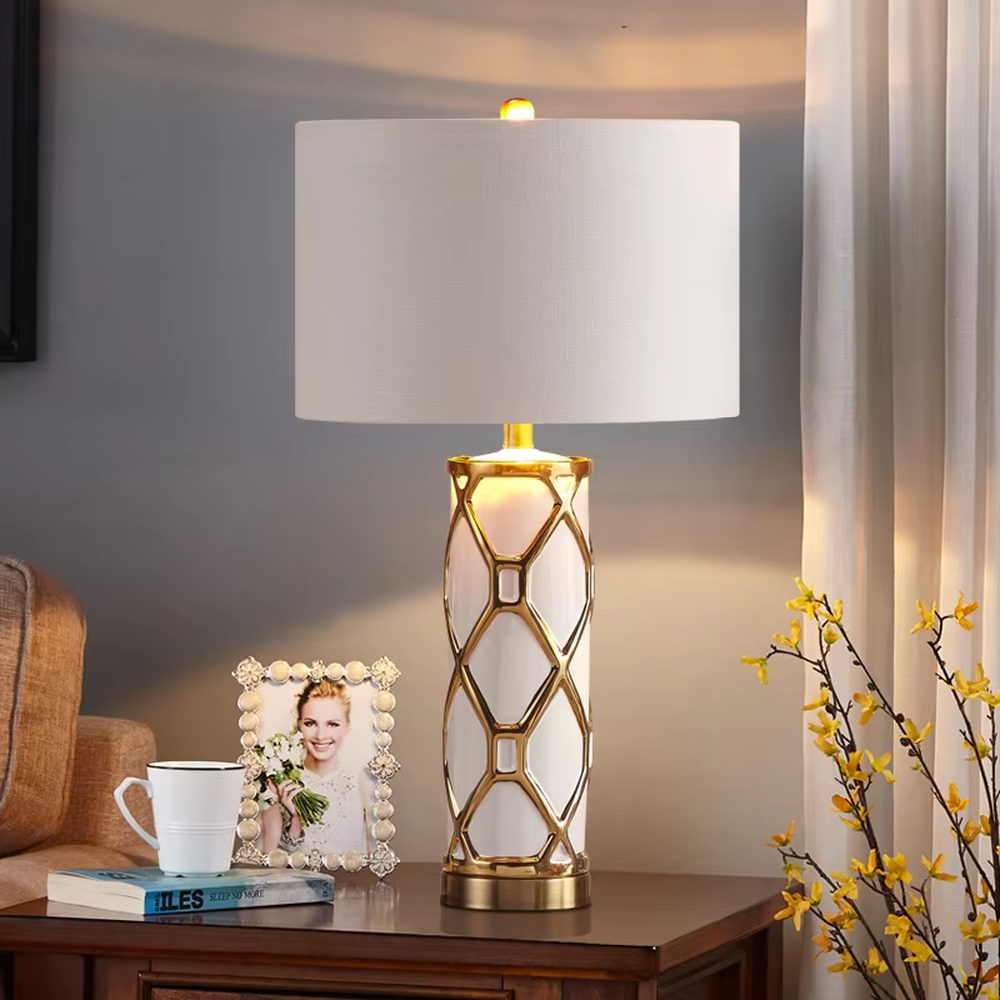 This is a luxury-style gold and white geometric hollow table lamp, with a gold geometric hollow base, white cylinder, and beige fabric circular lampshade, featuring an exquisite and elegant design with strong luxury texture. It is used for local desktop lighting and decoration, creating a luxury and warm atmosphere. Suitable for luxury-style home bedroom bedside tables, high-end hotel guest rooms, European-style living room side tables and other places.