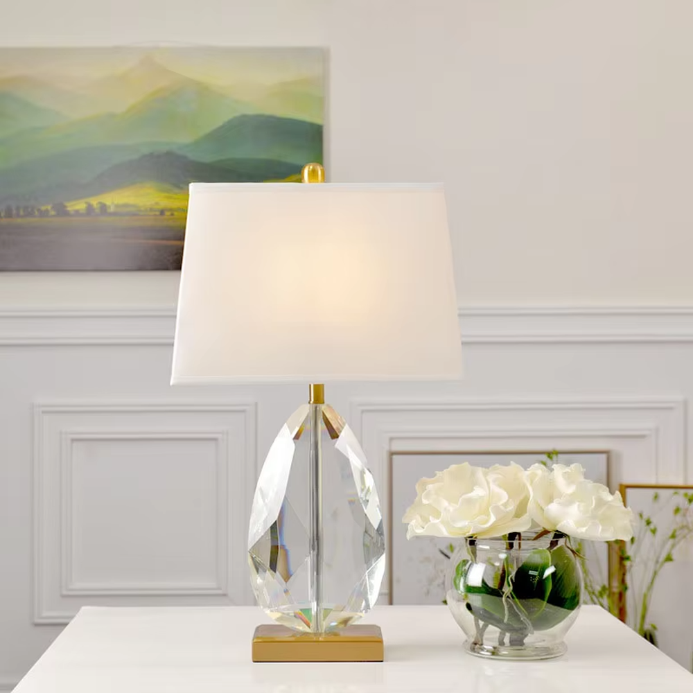 This is a light luxury exquisite water-drop shaped crystal table lamp, with a transparent cut water-drop crystal base paired with golden metal parts and a white square fabric lampshade. The design is simple and luxurious, with soft and elegant lighting. It is used for local desktop lighting and decoration, creating a light luxury exquisite atmosphere. Suitable for light luxury-style bedroom bedside tables, modern minimalist living room side tables, high-end hotel guest rooms and other places.