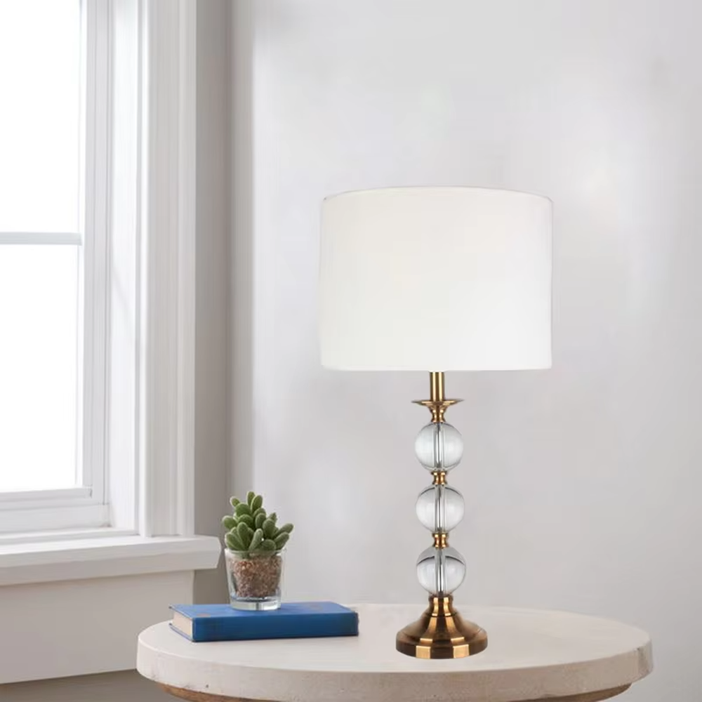 This is a light luxury exquisite crystal ball column table lamp, with a golden metal base and connectors, paired with transparent crystal balls and a white fabric round lampshade. It features a light luxury and exquisite design, with warm and soft lighting. It is used for local desktop lighting and decoration, creating a light luxury elegant atmosphere. Suitable for light luxury-style bedroom bedside tables, modern minimalist living room side tables, high-end hotel guest rooms and other places.