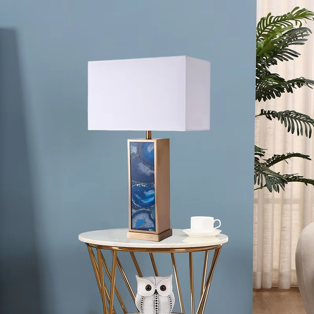 This is a luxury artistic agate-textured table lamp, with a golden metal frame inlaid with blue agate texture decoration and a white square fabric lampshade, featuring a minimalist and grand design with both artistic sense and luxury texture. It is used for local desktop lighting and decoration, creating a luxury artistic atmosphere. It is suitable for luxury-style home bedroom bedside tables, high-end hotel guest rooms, art exhibition hall display tables and other places.