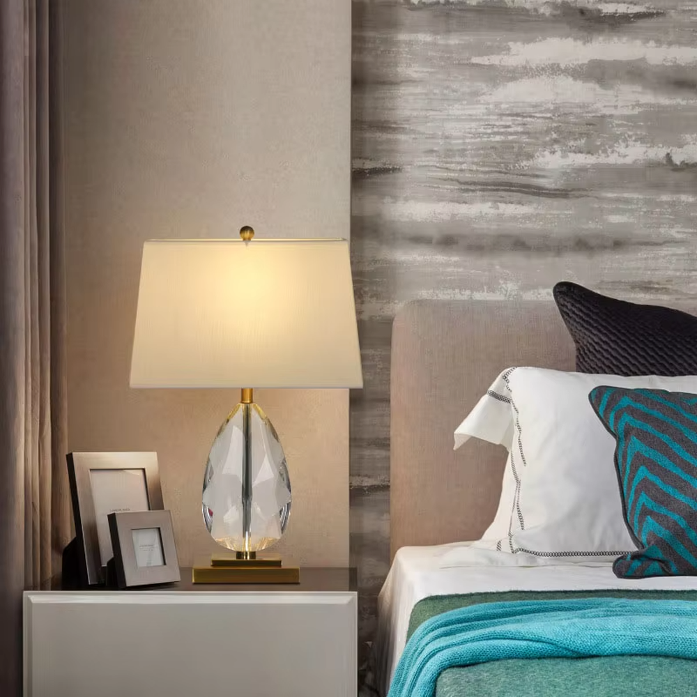 This is a light luxury exquisite water-drop shaped crystal table lamp, with a transparent cut water-drop crystal base paired with golden metal parts and a white square fabric lampshade. The design is simple and luxurious, with soft and elegant lighting. It is used for local desktop lighting and decoration, creating a light luxury exquisite atmosphere. Suitable for light luxury-style bedroom bedside tables, modern minimalist living room side tables, high-end hotel guest rooms and other places.