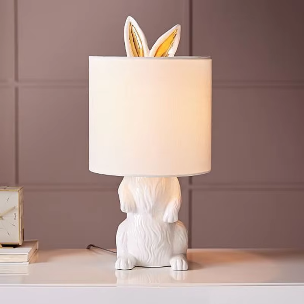 This is a cute and adorable rabbit-shaped table lamp, with a white rabbit-shaped base paired with golden ear decorations, and a white cylindrical lampshade. It has a warm and cute style, with soft and comfortable lighting.
