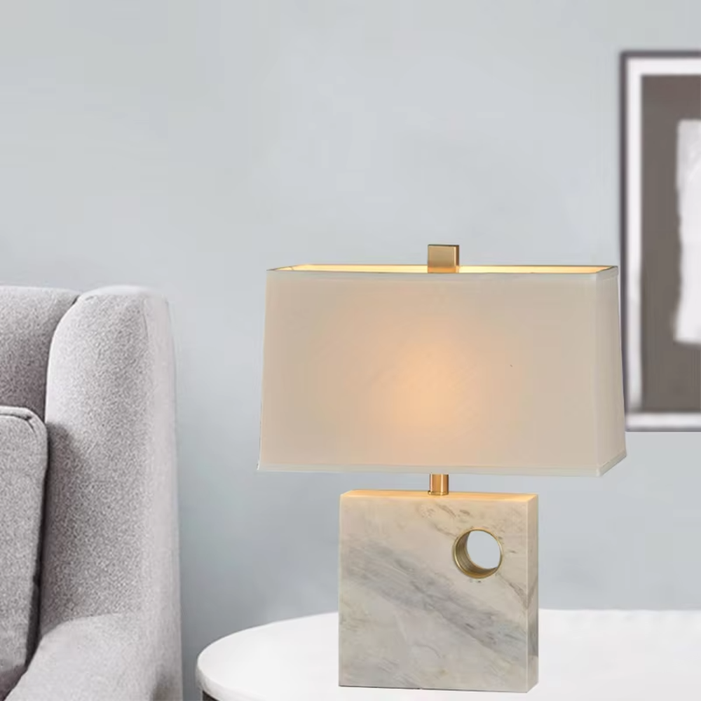 This is a light luxury elegant marble-based fabric table lamp, with a white marble base with golden ring decoration, paired with a beige fabric square lampshade and golden metal connectors. It features a minimalist light luxury design, with warm and soft lighting. It is used for local desktop lighting and decoration, creating a light luxury elegant atmosphere. Suitable for light luxury-style bedroom bedside tables, modern minimalist studies, high-end hotel guest rooms and other places.