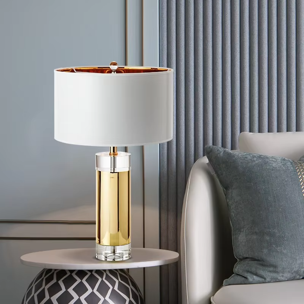 This is a luxury metal crystal table lamp, with a golden metal column, a transparent crystal base, and a white cylindrical fabric lampshade (with golden decoration on top), featuring a luxurious and elegant design with strong modern sense. It is used for local desktop lighting and decoration, creating a high-end luxury atmosphere. It is suitable for luxury-style home bedroom bedside tables, high-end hotel guest rooms, modern soft decoration display spaces and other places.