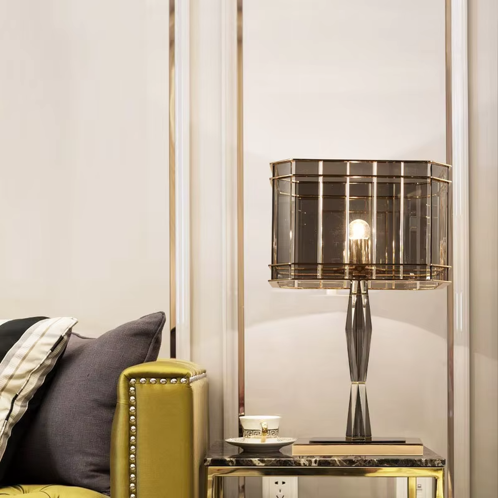This is a light luxury and luxurious smoky gray glass metal table lamp, with a smoky gray glass lampshade paired with a golden metal frame, a golden base, and a geometrically cut lamp post. It features an exquisite and grand design, with soft and textured lighting through the glass. It is used for local desktop lighting and decoration, creating a light luxury and luxurious atmosphere. Suitable for light luxury-style bedroom bedside tables, luxury living room side tables, high-end hotel guest rooms and other places.