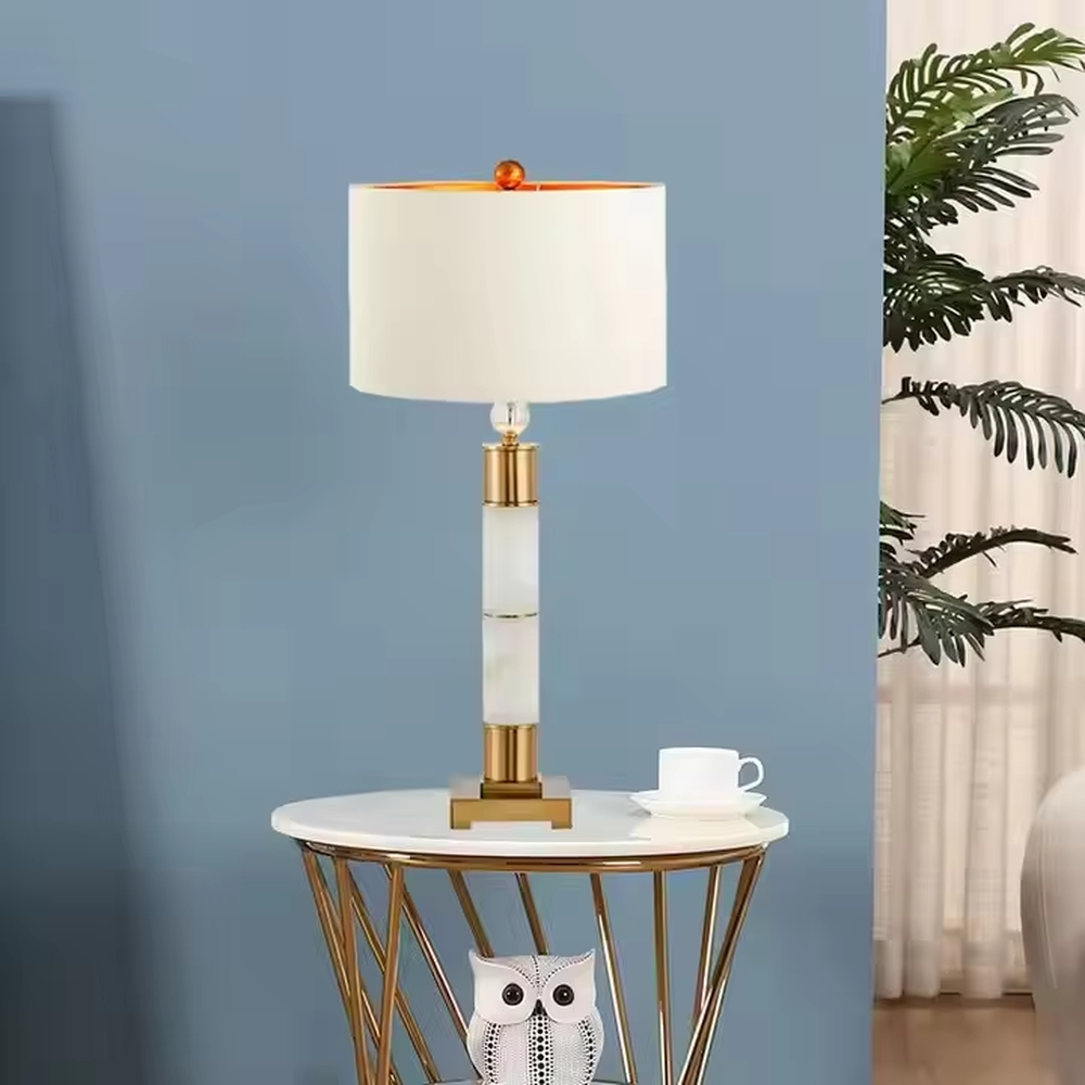 This is a luxury-style gold and white table lamp, combining golden metal and white cylinder, with a beige fabric circular lampshade, featuring a minimalist and grand design with strong luxury texture. It is used for local desktop lighting and decoration, creating a luxury and warm atmosphere. It is suitable for luxury-style home bedroom bedside tables, high-end hotel guest rooms, modern minimalist living room side tables and other places.
