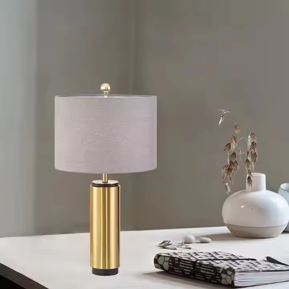 This is a light luxury minimalist golden cylinder table lamp, with a golden metal cylinder base paired with a light purple textured fabric lampshade, decorated with a small golden ball on top. The lighting is soft and warm, creating an elegant light luxury atmosphere.