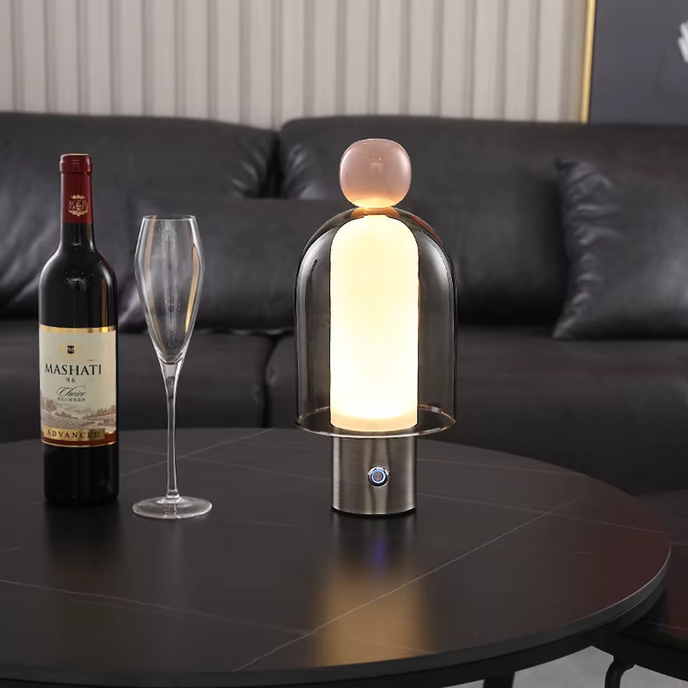 This is an artistic doll-style glass and metal table lamp, with a transparent glass shell, pink top, and metal base. Its unique shape is like an artistic doll, featuring a strong modern artistic sense. It is used for local desktop lighting and decoration, creating an artistic and modern atmosphere. Suitable for art exhibition halls, corners of modern home studies, designer studios and other places.