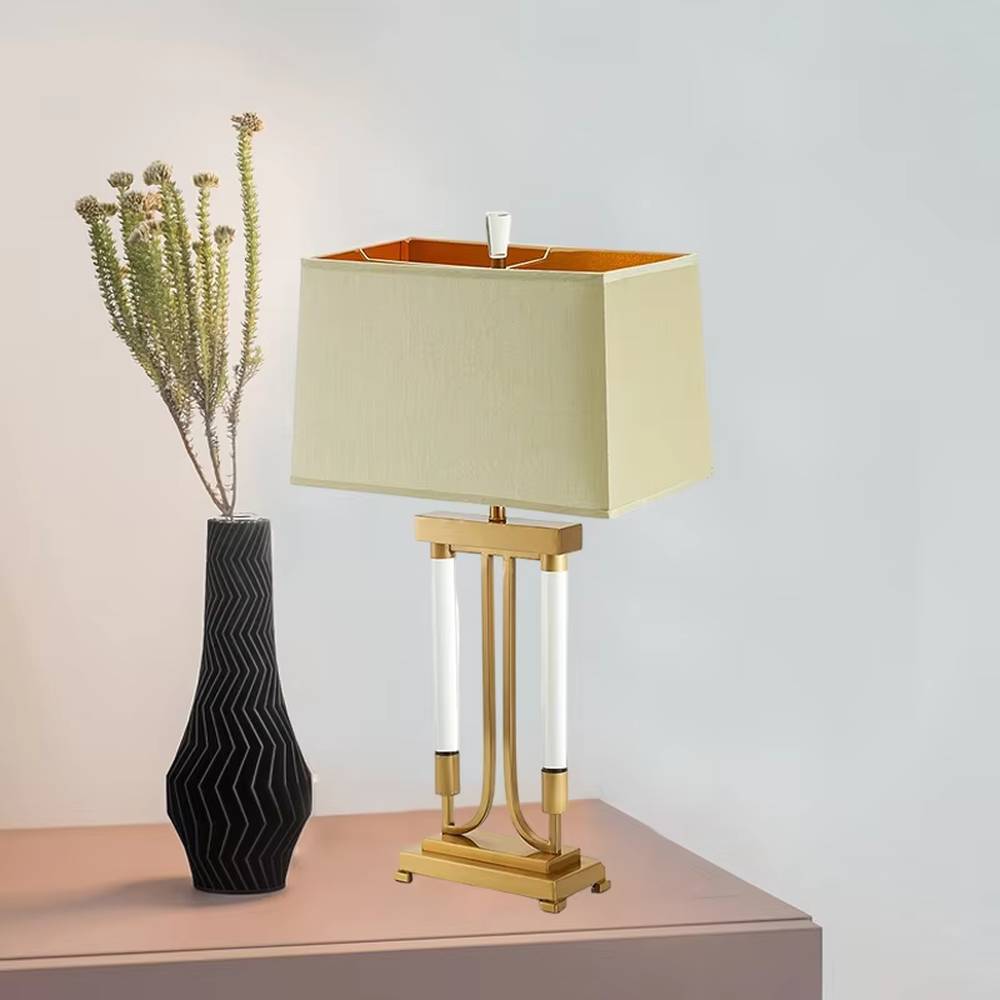 This is a retro elegant-style gold and white fabric table lamp, with a golden metal bracket (with white column elements) and a beige fabric square lampshade (orange interior). It features a retro and exquisite design, with warm and soft lighting. It is used for local desktop lighting and decoration, creating a retro and elegant atmosphere. Suitable for retro-style bedrooms, European-style living rooms, high-end homestays and other places.