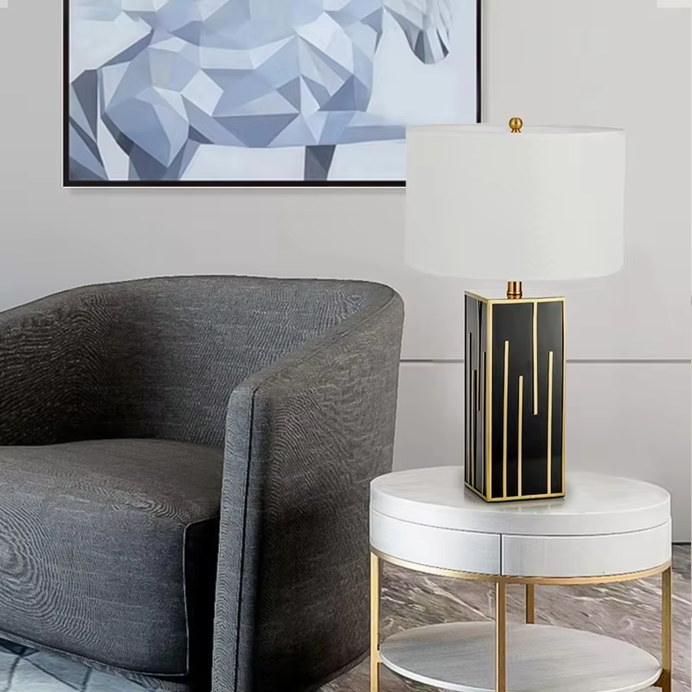 This is a luxury geometric - shaped table lamp with a black - gold geometric base (decorated with gold lines) and a white cylindrical fabric lampshade, featuring a minimalist luxury design with strong modern artistic sense. It is used for local desktop lighting and decoration, creating a luxurious and elegant atmosphere. It is suitable for luxury - style home bedroom bedside tables, high - end hotel guest rooms, modern soft decoration display spaces and other places.