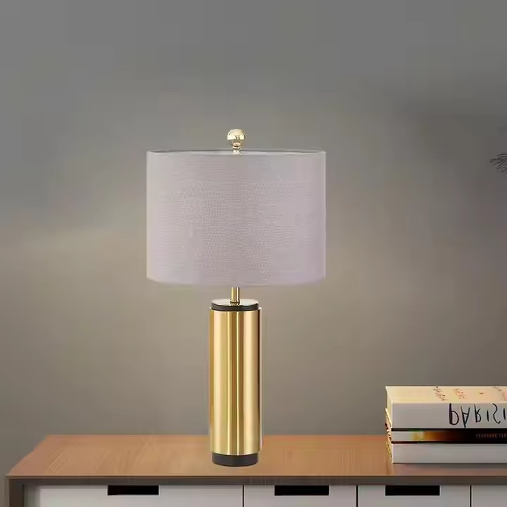 This is a light luxury minimalist golden cylinder table lamp, with a golden metal cylinder base paired with a light purple textured fabric lampshade, decorated with a small golden ball on top. The lighting is soft and warm, creating an elegant light luxury atmosphere.
