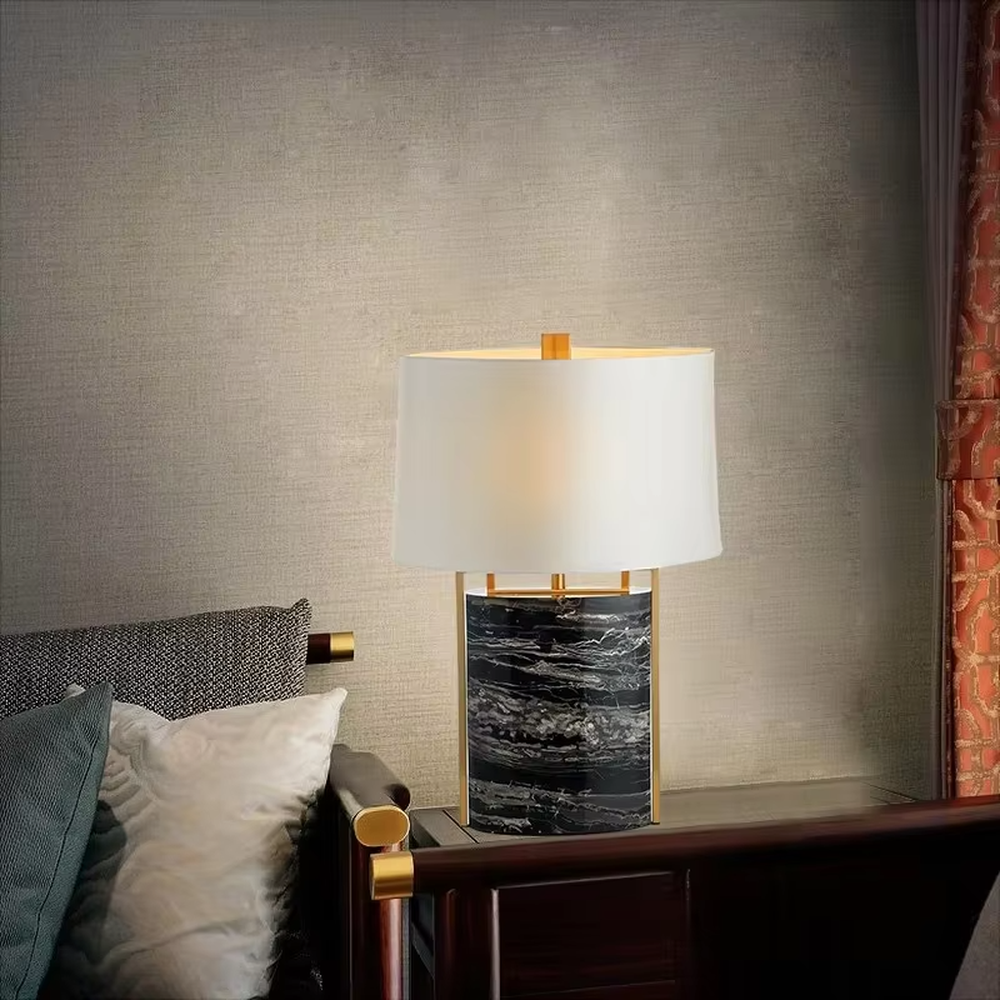 This is a luxury marble table lamp, with a black-veined marble base, a golden metal frame, and a white cylindrical fabric lampshade, featuring a minimalist luxury design with strong modern texture. It is used for local desktop lighting and decoration, creating a high-end luxury atmosphere. It is suitable for luxury-style home bedroom bedside tables, high-end hotel guest rooms, modern soft decoration display spaces and other places.