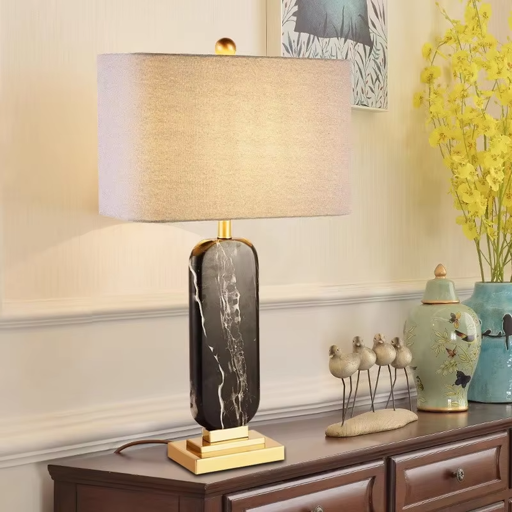 This is a luxury black-gold marble table lamp, with a black-veined marble base, golden metal components, and a gray fabric square lampshade, featuring a minimalist luxury design with high-quality texture. It is used for local desktop lighting and decoration, creating a high-end luxury atmosphere. It is suitable for luxury-style home bedroom bedside tables, high-end hotel guest rooms, modern soft decoration display spaces and other places.
