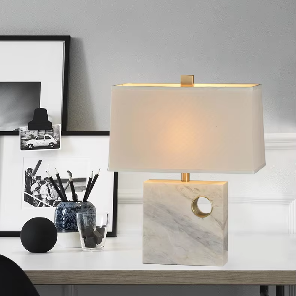 This is a light luxury elegant marble-based fabric table lamp, with a white marble base with golden ring decoration, paired with a beige fabric square lampshade and golden metal connectors. It features a minimalist light luxury design, with warm and soft lighting. It is used for local desktop lighting and decoration, creating a light luxury elegant atmosphere. Suitable for light luxury-style bedroom bedside tables, modern minimalist studies, high-end hotel guest rooms and other places.