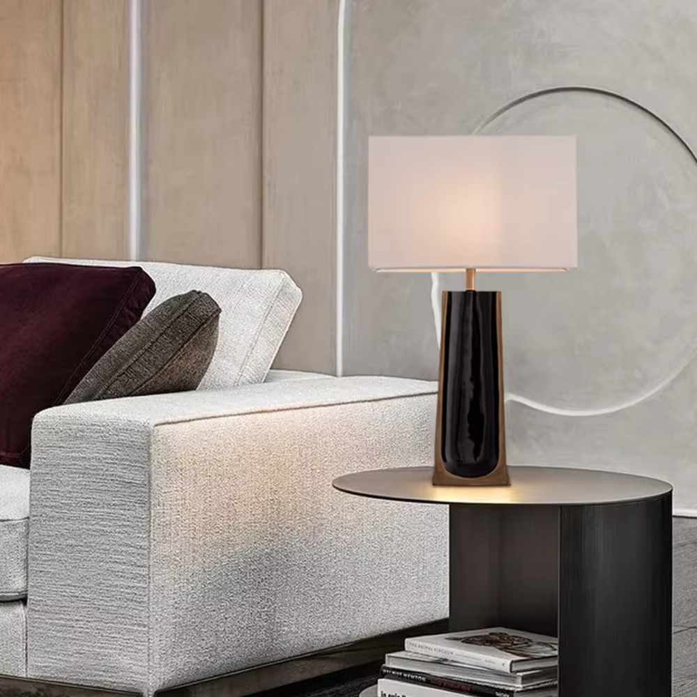 This is a light luxury modern-style black and gold column fabric table lamp, with a black and gold column base (with golden decorative edges) paired with a white fabric square lampshade. It features a minimalist light luxury design, with warm and soft lighting. It is used for local desktop lighting and decoration, creating a light luxury elegant atmosphere. Suitable for light luxury-style bedroom bedside tables, modern minimalist living room side tables, high-end hotel guest rooms and other places.