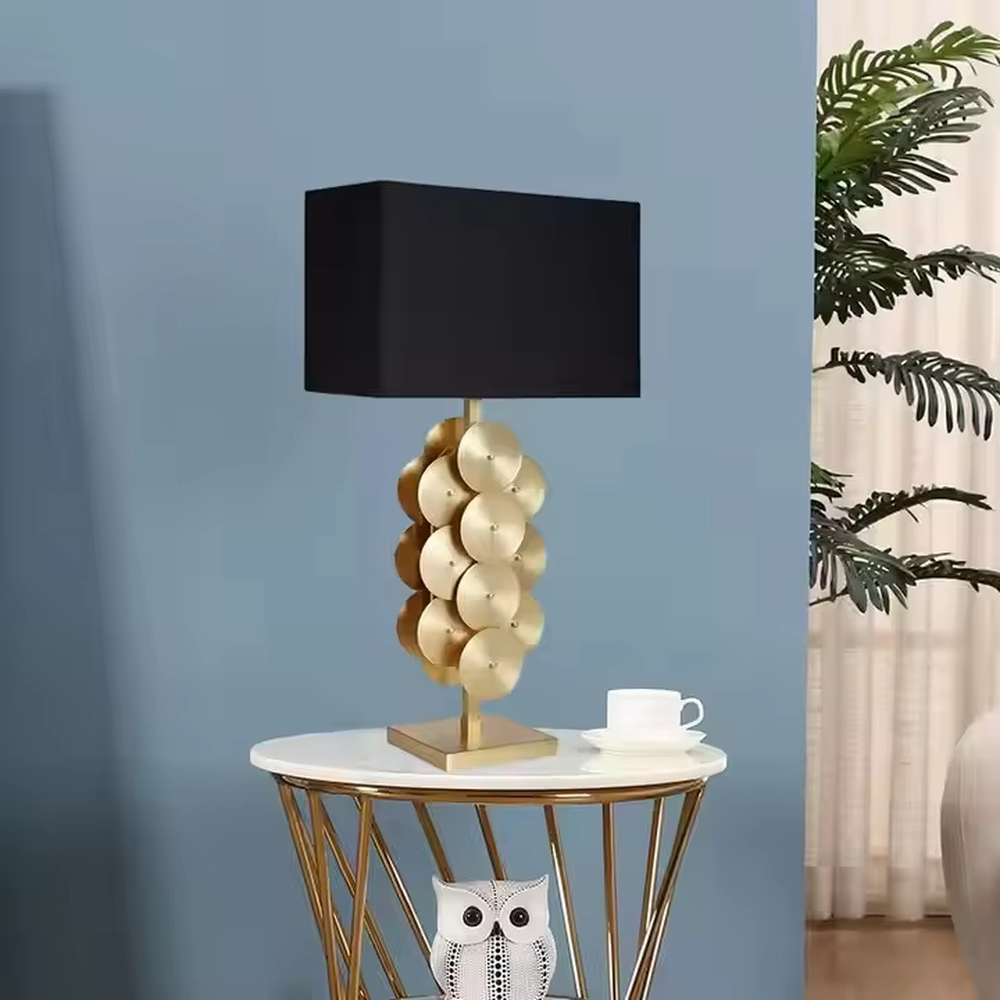 This is a luxury-style black and gold table lamp, with a golden disc-stacked base and a black square fabric lampshade, featuring a minimalist and grand design with strong luxury texture. It is used for local desktop lighting and decoration, creating a luxury and modern atmosphere. It is suitable for luxury-style home bedroom bedside tables, high-end hotel guest rooms, modern minimalist living room side tables and other places.