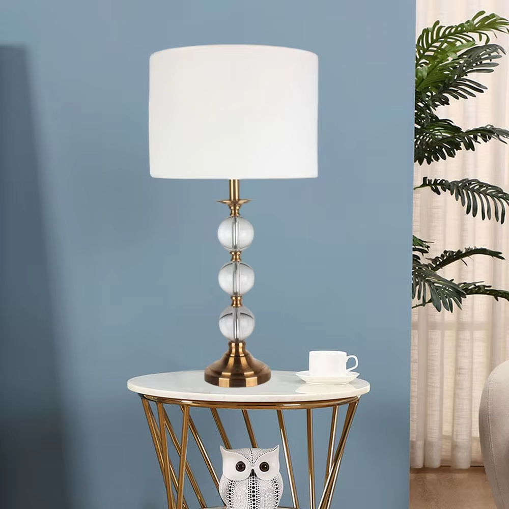 This is a light luxury exquisite crystal ball column table lamp, with a golden metal base and connectors, paired with transparent crystal balls and a white fabric round lampshade. It features a light luxury and exquisite design, with warm and soft lighting. It is used for local desktop lighting and decoration, creating a light luxury elegant atmosphere. Suitable for light luxury-style bedroom bedside tables, modern minimalist living room side tables, high-end hotel guest rooms and other places.
