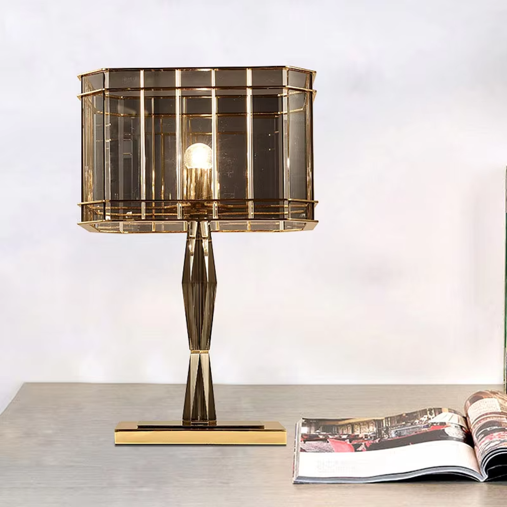 This is a light luxury and luxurious smoky gray glass metal table lamp, with a smoky gray glass lampshade paired with a golden metal frame, a golden base, and a geometrically cut lamp post. It features an exquisite and grand design, with soft and textured lighting through the glass. It is used for local desktop lighting and decoration, creating a light luxury and luxurious atmosphere. Suitable for light luxury-style bedroom bedside tables, luxury living room side tables, high-end hotel guest rooms and other places.