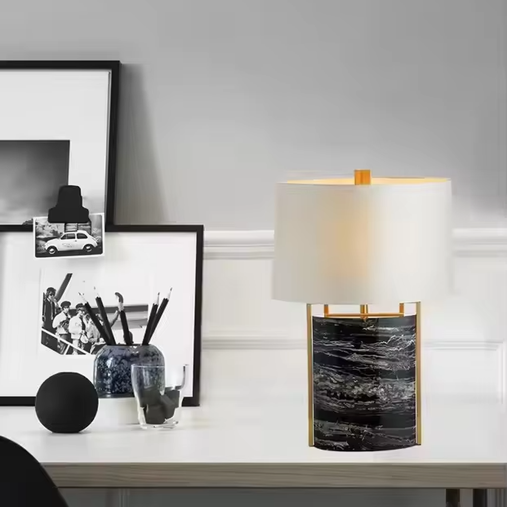 This is a luxury marble table lamp, with a black-veined marble base, a golden metal frame, and a white cylindrical fabric lampshade, featuring a minimalist luxury design with strong modern texture. It is used for local desktop lighting and decoration, creating a high-end luxury atmosphere. It is suitable for luxury-style home bedroom bedside tables, high-end hotel guest rooms, modern soft decoration display spaces and other places.
