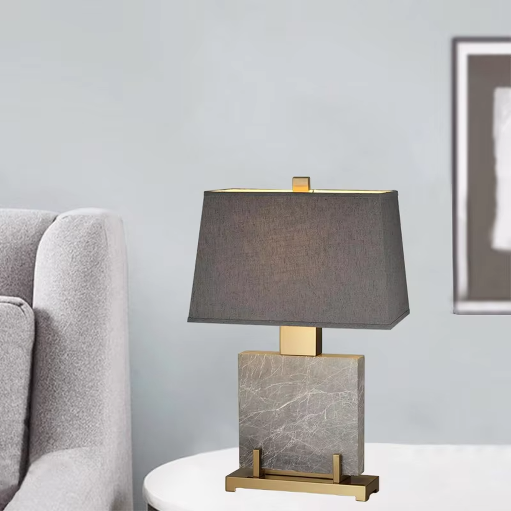This is a light luxury modern-style marble metal fabric table lamp, with a gray marble base paired with golden metal brackets and connectors, and a gray fabric square lampshade. It features a minimalist and grand design with high texture. It is used for local desktop lighting and decoration, creating a light luxury and elegant atmosphere. Suitable for light luxury-style bedroom bedside tables, modern minimalist living room side tables, high-end hotel guest rooms and other places.