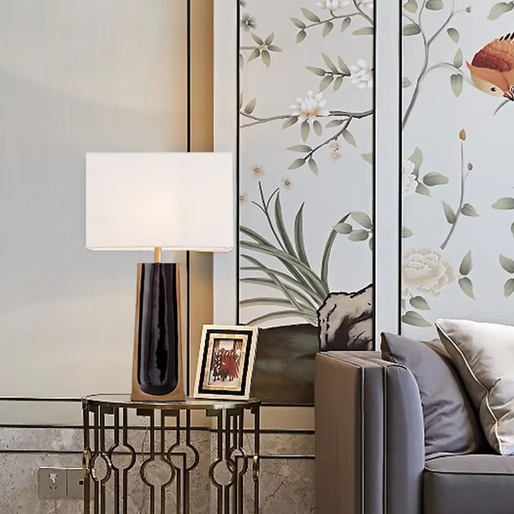 This is a light luxury modern-style black and gold column fabric table lamp, with a black and gold column base (with golden decorative edges) paired with a white fabric square lampshade. It features a minimalist light luxury design, with warm and soft lighting. It is used for local desktop lighting and decoration, creating a light luxury elegant atmosphere. Suitable for light luxury-style bedroom bedside tables, modern minimalist living room side tables, high-end hotel guest rooms and other places.