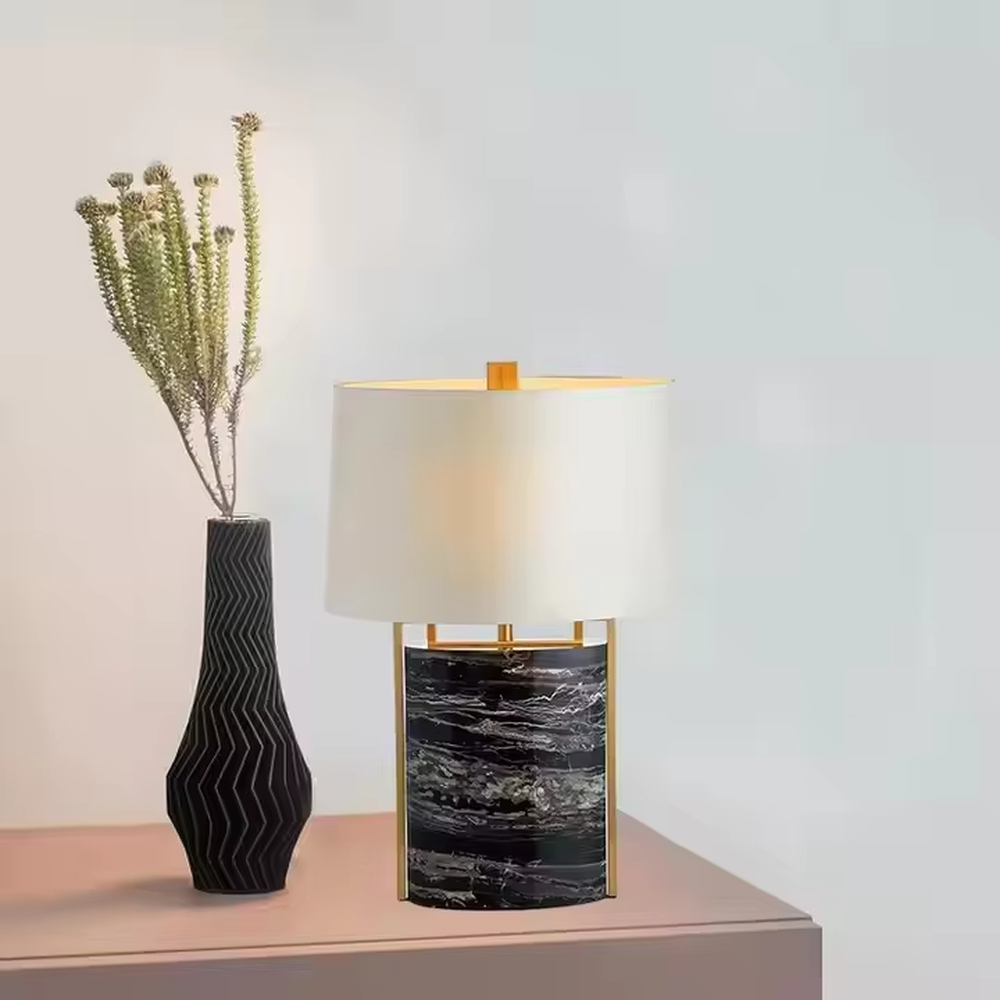 This is a luxury marble table lamp, with a black-veined marble base, a golden metal frame, and a white cylindrical fabric lampshade, featuring a minimalist luxury design with strong modern texture. It is used for local desktop lighting and decoration, creating a high-end luxury atmosphere. It is suitable for luxury-style home bedroom bedside tables, high-end hotel guest rooms, modern soft decoration display spaces and other places.