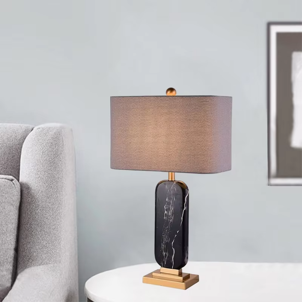 This is a luxury black-gold marble table lamp, with a black-veined marble base, golden metal components, and a gray fabric square lampshade, featuring a minimalist luxury design with high-quality texture. It is used for local desktop lighting and decoration, creating a high-end luxury atmosphere. It is suitable for luxury-style home bedroom bedside tables, high-end hotel guest rooms, modern soft decoration display spaces and other places.