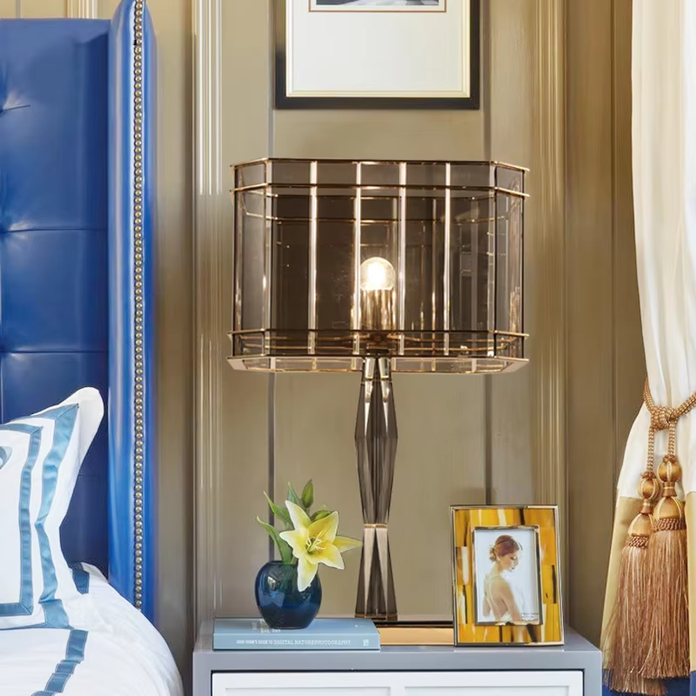 This is a light luxury and luxurious smoky gray glass metal table lamp, with a smoky gray glass lampshade paired with a golden metal frame, a golden base, and a geometrically cut lamp post. It features an exquisite and grand design, with soft and textured lighting through the glass. It is used for local desktop lighting and decoration, creating a light luxury and luxurious atmosphere. Suitable for light luxury-style bedroom bedside tables, luxury living room side tables, high-end hotel guest rooms and other places.