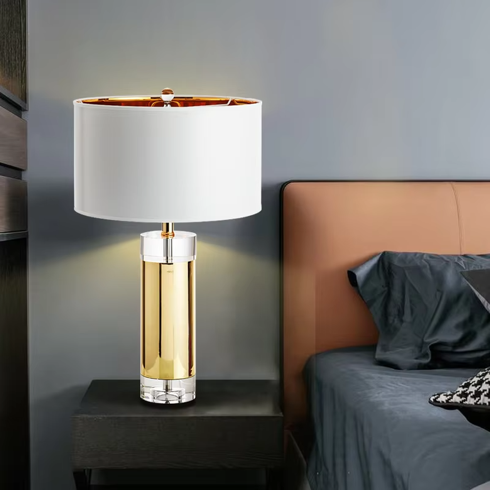 This is a luxury metal crystal table lamp, with a golden metal column, a transparent crystal base, and a white cylindrical fabric lampshade (with golden decoration on top), featuring a luxurious and elegant design with strong modern sense. It is used for local desktop lighting and decoration, creating a high-end luxury atmosphere. It is suitable for luxury-style home bedroom bedside tables, high-end hotel guest rooms, modern soft decoration display spaces and other places.