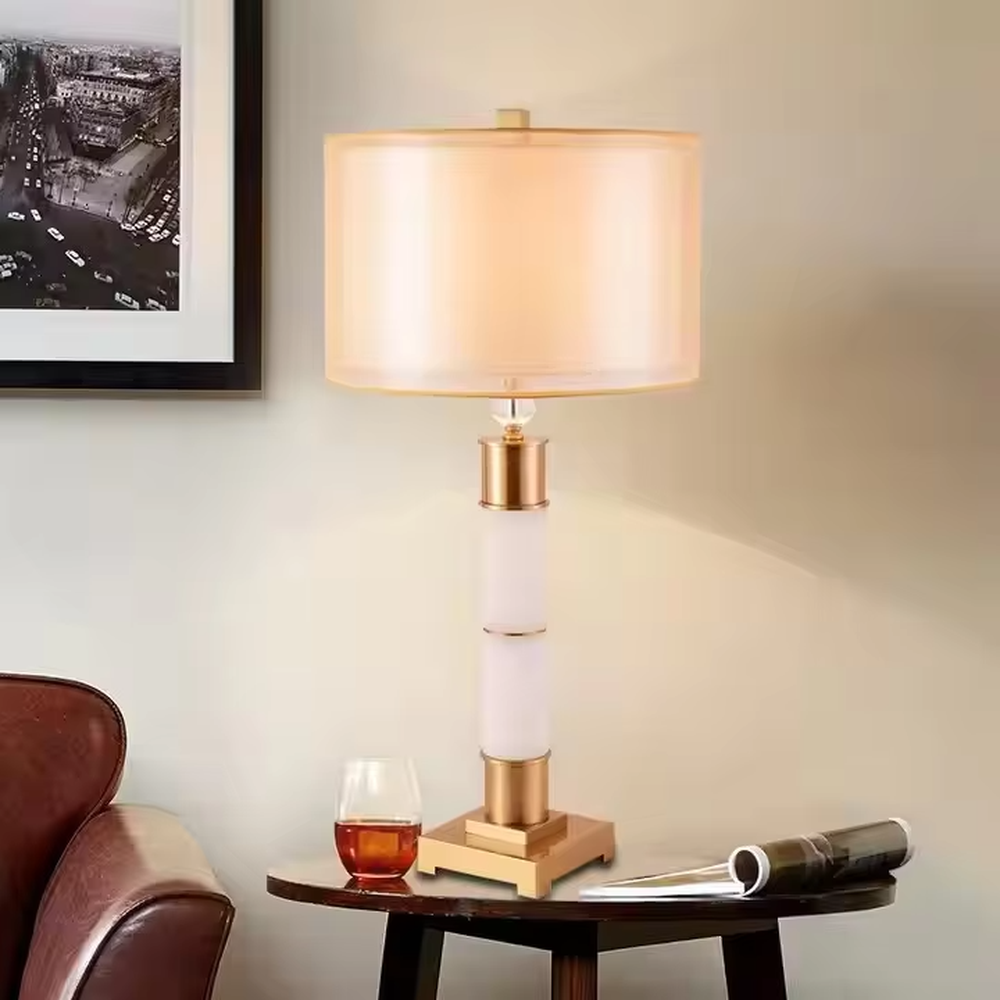 This is a luxury-style gold and white table lamp, combining golden metal and white cylinder, with a beige fabric circular lampshade, featuring a minimalist and grand design with strong luxury texture. It is used for local desktop lighting and decoration, creating a luxury and warm atmosphere. It is suitable for luxury-style home bedroom bedside tables, high-end hotel guest rooms, modern minimalist living room side tables and other places.
