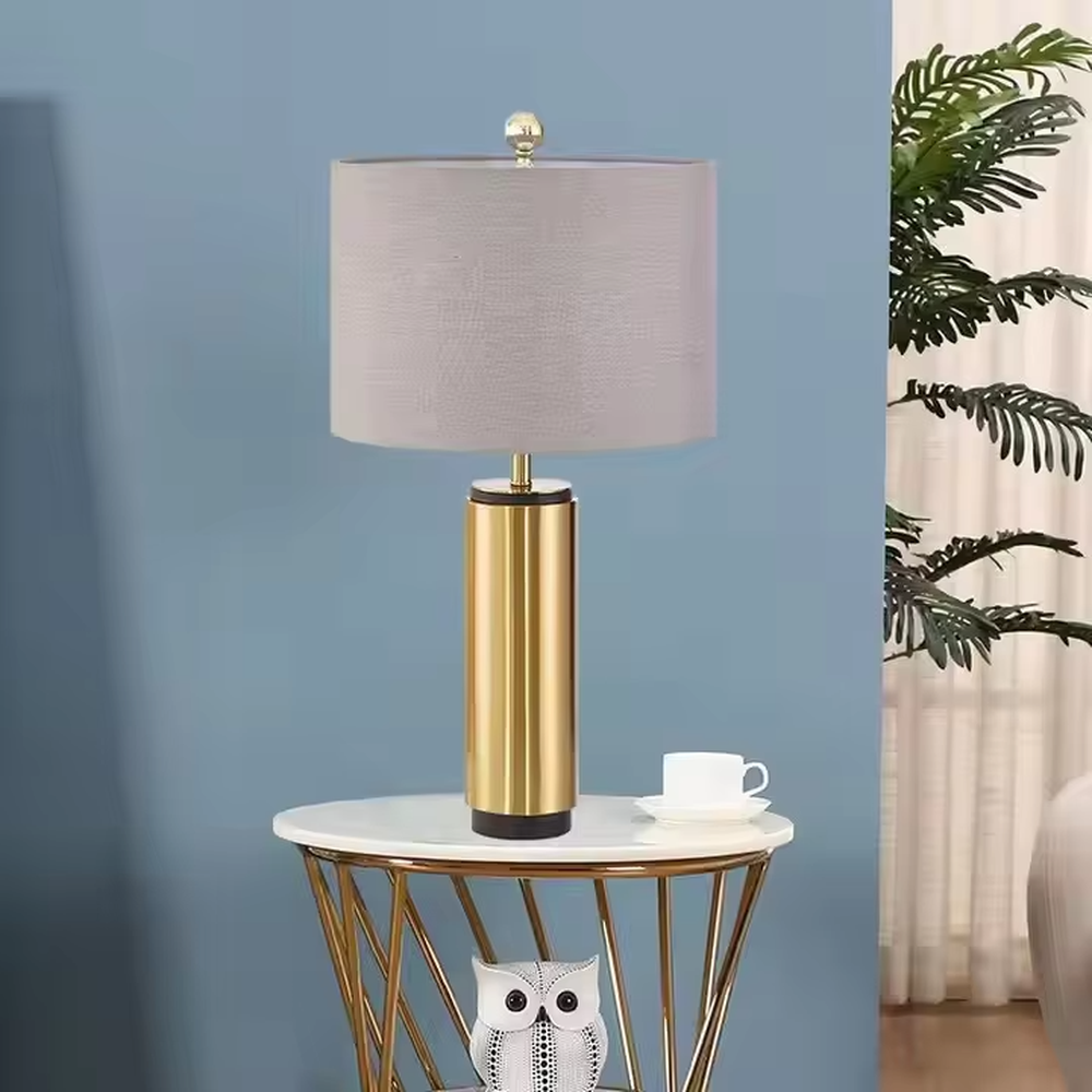 This is a light luxury minimalist golden cylinder table lamp, with a golden metal cylinder base paired with a light purple textured fabric lampshade, decorated with a small golden ball on top. The lighting is soft and warm, creating an elegant light luxury atmosphere.
